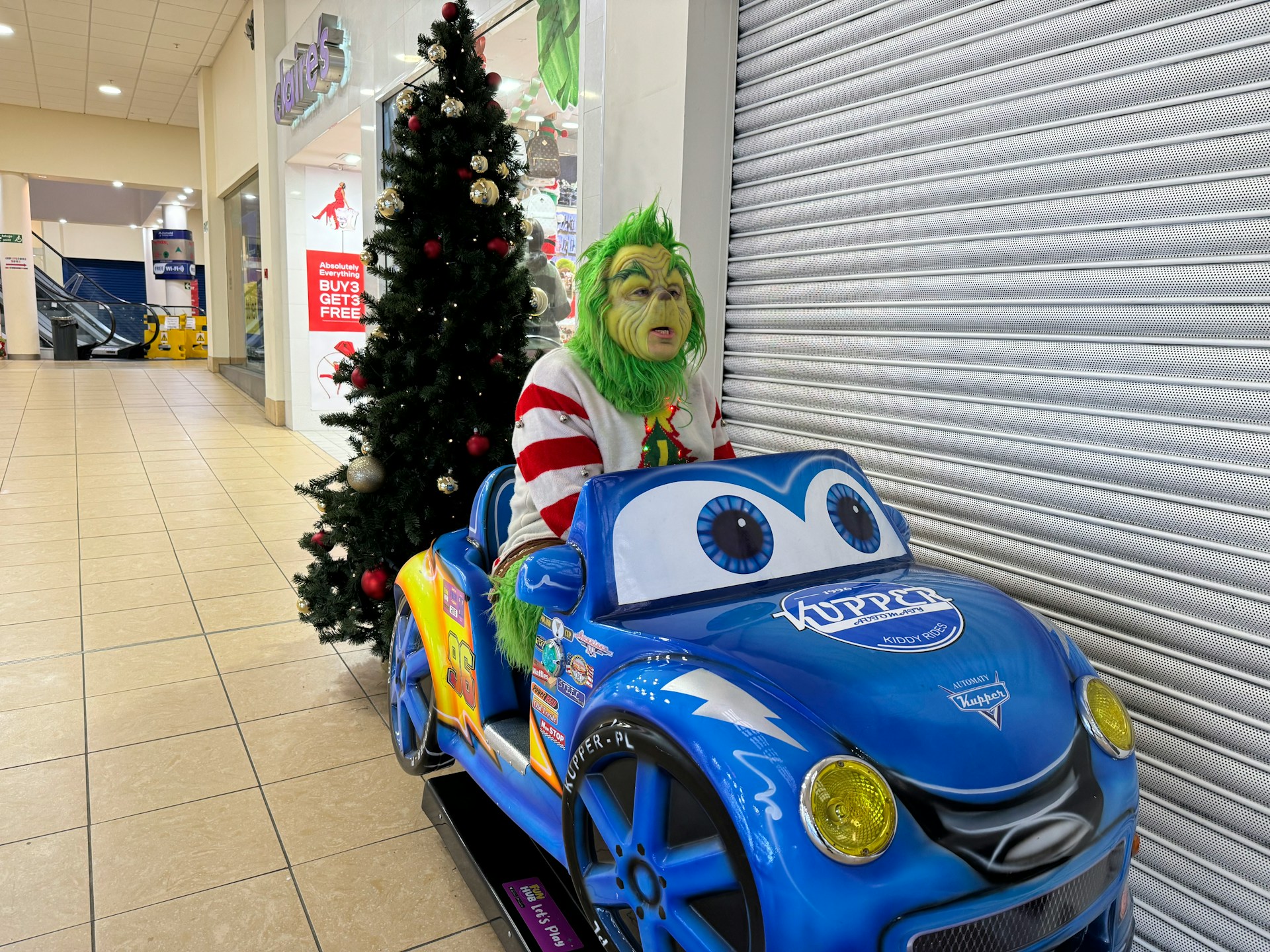 A picture of The Grinch in a kid's ride-on car in a mall. Filming yourself doing this is a great example of what not to do online at Christmas!